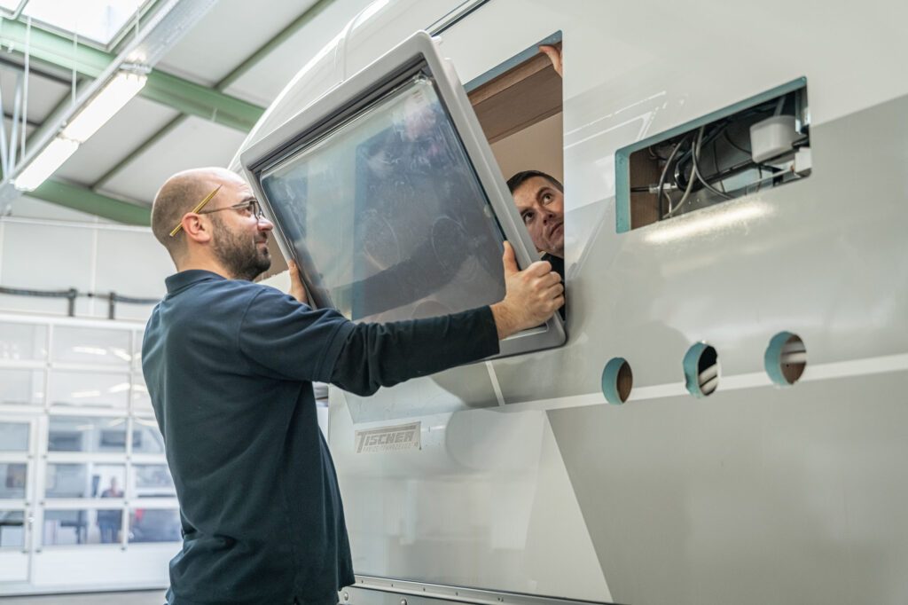 Zwei Mitarbeiter der Tischer Manufaktur setzen gemeinsam ein Fenster in die Seitenwand einer Wohnkabine ein.