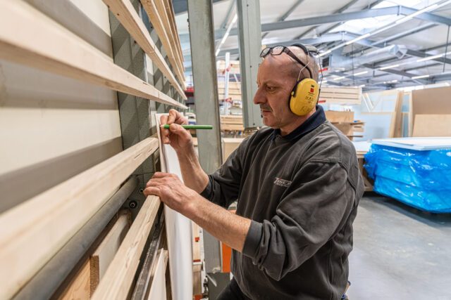 Ein Mitarbeiter greift im Materiallager der Tischer Manufaktur nach einer Holzleiste – Gehörschutz und Maßarbeit inklusive.