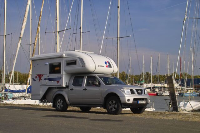2014_Tischer_Historie_95 - Tischer Wohnkabine auf grauem Pick-up – geparkt vor Hafen mit Segelbooten.