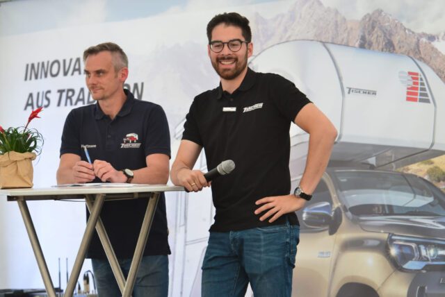 Zwei Tischer-Mitarbeiter auf der Bühne während des Treffens – einer spricht ins Mikrofon, der andere lächelt.