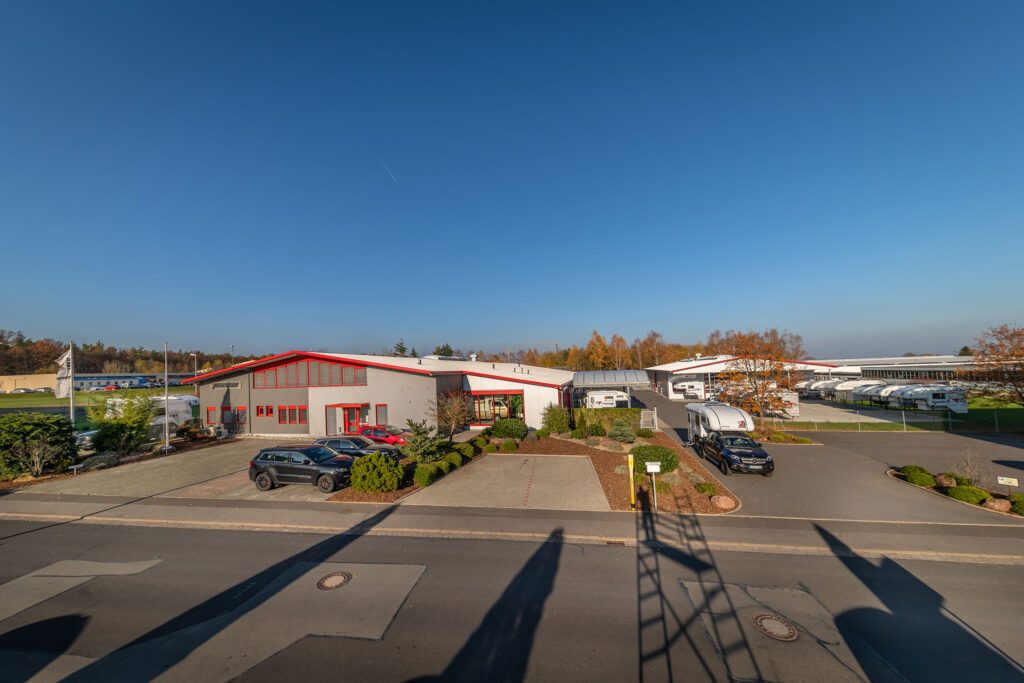 Außenaufnahme des Tischer-Werksgeländes mit Verwaltungsgebäude, geparkten Fahrzeugen und mehreren Kabinen.