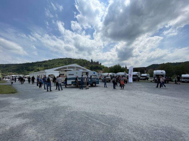 Veranstaltungsbereich vor dem Tischer-Werk mit Teilnehmern und abgestellten Fahrzeugen unter wolkigem Himmel.