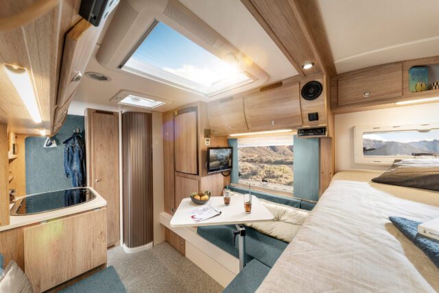 Tischer Wohnkabine - Wohnbereich mit Bett und Sitzgruppe in heller Holzoptik – Panoramafenster mit Blick auf Gebirgslandschaft.