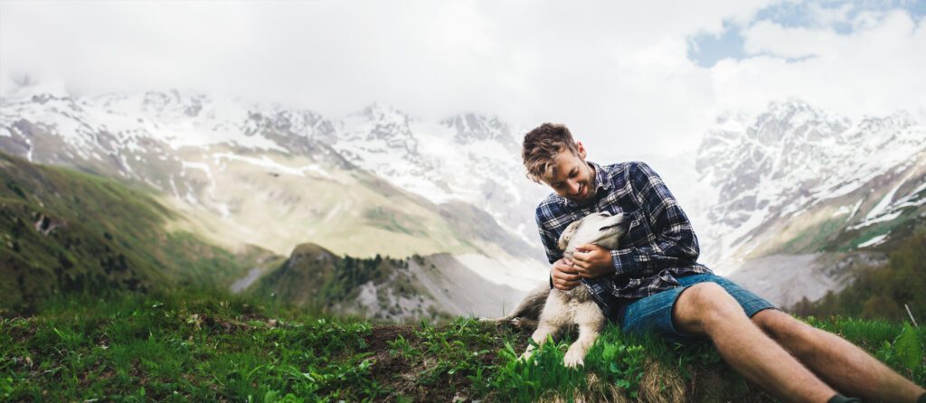 Young Caucasian man sitting on the grass with dog on the background of Caucasus mountains
