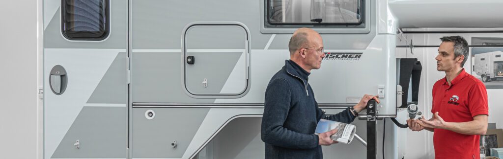 Zwei Mönner vor der Seitenwand einer Tischer-Wohnkabine mit klar erkennbarer Tür und grafischen Elementen.